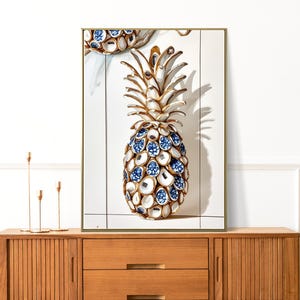 Puede incluir: Una pintura representa una piña dorada y blanca con detalles azules y blancos. La piña es el punto focal de la obra de arte, que está enmarcada y expuesta en una pared.