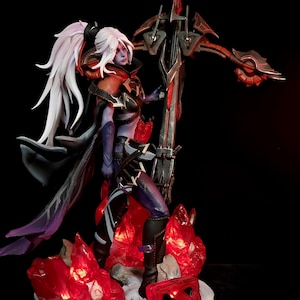 Puede incluir: Figura detallada de un personaje femenino con cabello blanco, que lleva una armadura oscura y sostiene un arma grande. La figura se encuentra sobre una base con detalles rojos brillantes y un logotipo rojo. El diseño general tiene un tema de fantasía.