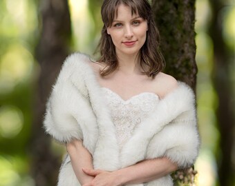 May include: A white faux fur shawl with a soft, fluffy texture. The shawl is draped over the shoulders of a person wearing a white lace dress.