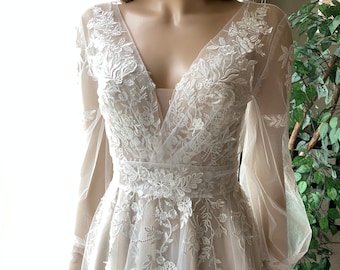 May include: A white lace wedding dress with long sleeves and a plunging V-neckline. The dress has a fitted bodice and a flowing skirt.