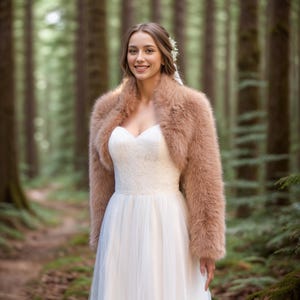 May include: A woman wearing a white wedding dress and a light brown faux fur shrug. The dress has a sweetheart neckline and a tulle skirt. The background is a forest with tall trees.