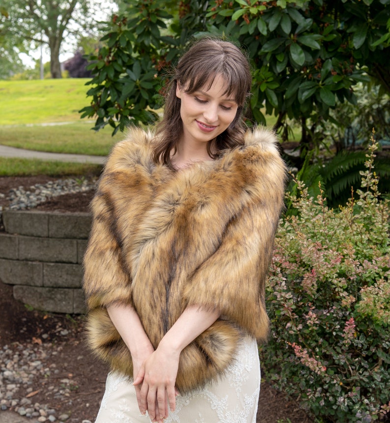 May include: A faux fur shawl in a brown and tan color with a soft, fluffy texture. The shawl is draped over the shoulders of a person wearing a white dress.