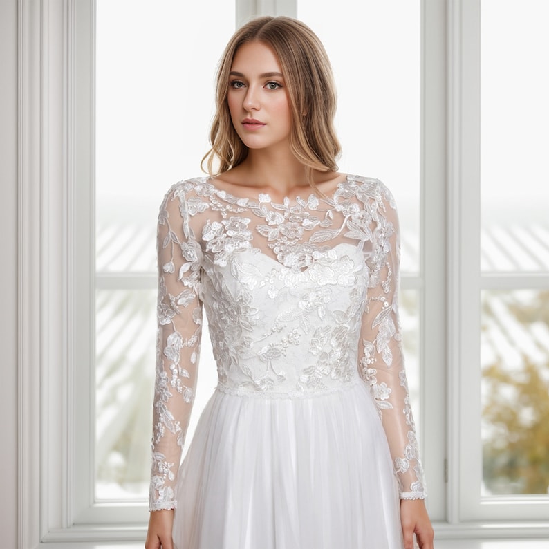 Op de afbeelding: Een witte trouwjurk met een doorschijnende, lange mouwen kanten overlay. De overlay heeft bloemenborduurwerk over het lijfje en de mouwen. De jurk heeft een sweetheart halslijn en een vloeiende tule rok.