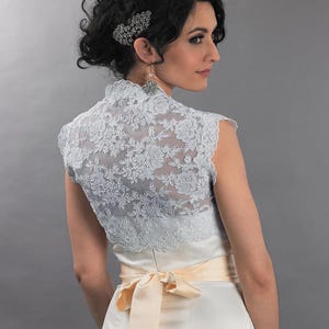 May include: A light blue lace bolero with a high collar, worn over a white satin dress with a peach-colored sash tied in a bow. The model has dark hair styled up with a decorative hairpiece and earrings. The background is a solid gray.