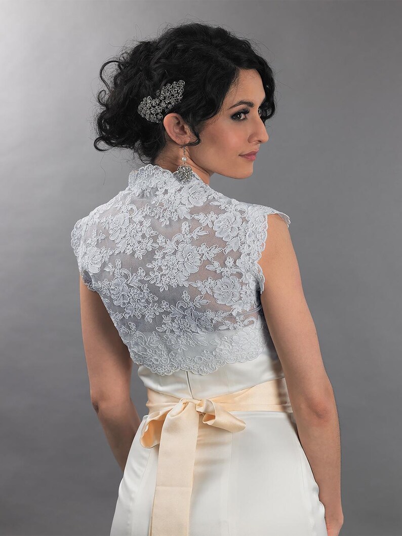 May include: A light blue lace bolero with a high collar, worn over a white satin dress with a peach-colored sash tied in a bow. The model has dark hair styled up with a decorative hairpiece and earrings. The background is a solid gray.