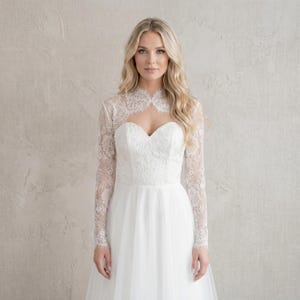 May include: A white lace bridal bolero with long sleeves, worn over a white wedding dress. The bolero features a delicate floral lace pattern and a high neckline. The dress has a sweetheart neckline and a flowing tulle skirt.