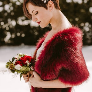 May include: A bride wearing a white dress with a red faux fur shawl. She is holding a bouquet of red and white flowers. The bride has short, dark hair with a silver hairpiece.