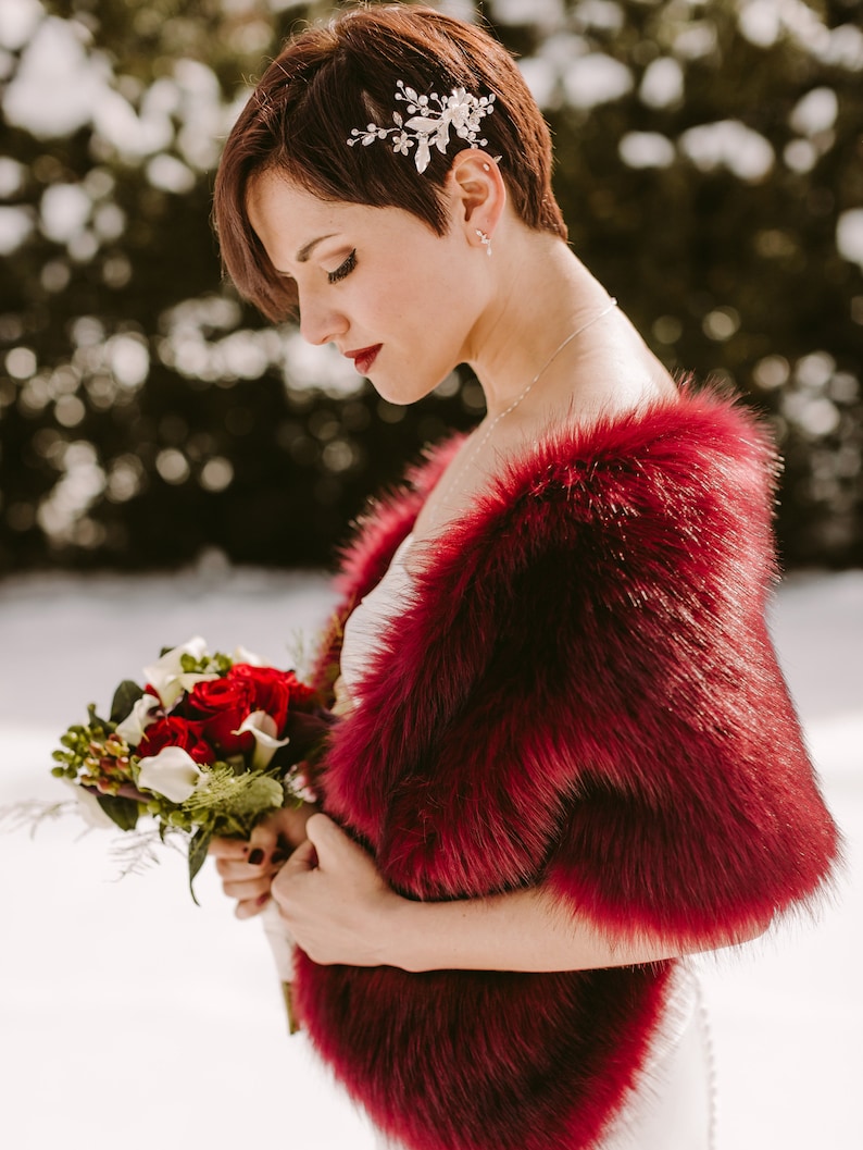 May include: A bride wearing a white dress with a red faux fur shawl. She is holding a bouquet of red and white flowers. The bride has short, dark hair with a silver hairpiece.