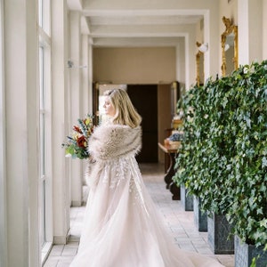 May include: A bride in a white wedding dress with a fur shawl, holding a bouquet of red, green, and blue flowers. She is standing in a hallway with white walls and a green ivy wall on the right.