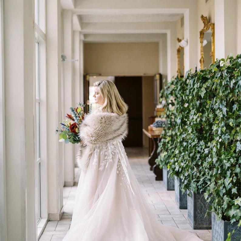 May include: A bride in a white wedding dress with a fur shawl, holding a bouquet of red, green, and blue flowers. She is standing in a hallway with white walls and a green ivy wall on the right.