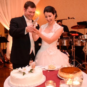 May include: A bride and groom cutting a white wedding cake with a silver knife. The bride is wearing a white lace dress and the groom is wearing a black suit. There is a band playing in the background.