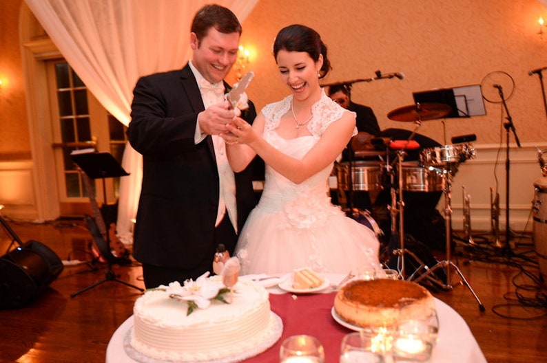 May include: A bride and groom cutting a white wedding cake with a silver knife. The bride is wearing a white lace dress and the groom is wearing a black suit. There is a band playing in the background.