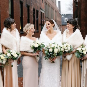 May include: A group of six women wearing white faux fur wraps and holding bouquets of white roses and greenery. The women are wearing long, flowing dresses in shades of beige and cream. The bride is in the center, wearing a white lace wedding dress.