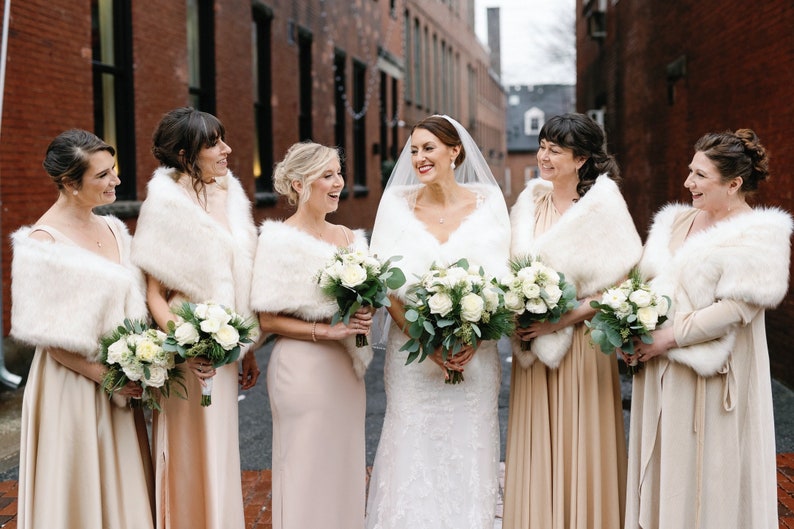 May include: A group of six women wearing white faux fur wraps and holding bouquets of white roses and greenery. The women are wearing long, flowing dresses in shades of beige and cream. The bride is in the center, wearing a white lace wedding dress.