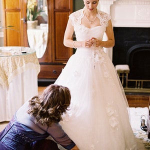 May include: A bride in a white wedding dress with a lace overlay and a pearl necklace is having her shoes fastened by a woman in a dark blue dress.