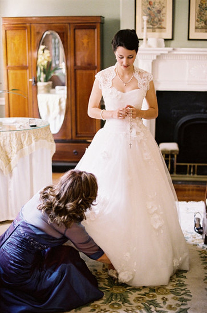 May include: A bride in a white wedding dress with a lace overlay and a pearl necklace is having her shoes fastened by a woman in a dark blue dress.