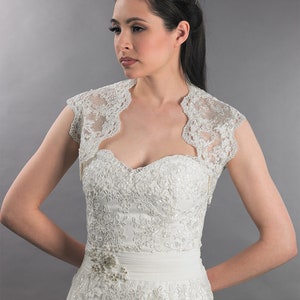 May include: A white lace bridal bolero with short sleeves and a scoop neckline. The bolero is made of delicate lace and has a scalloped edge.