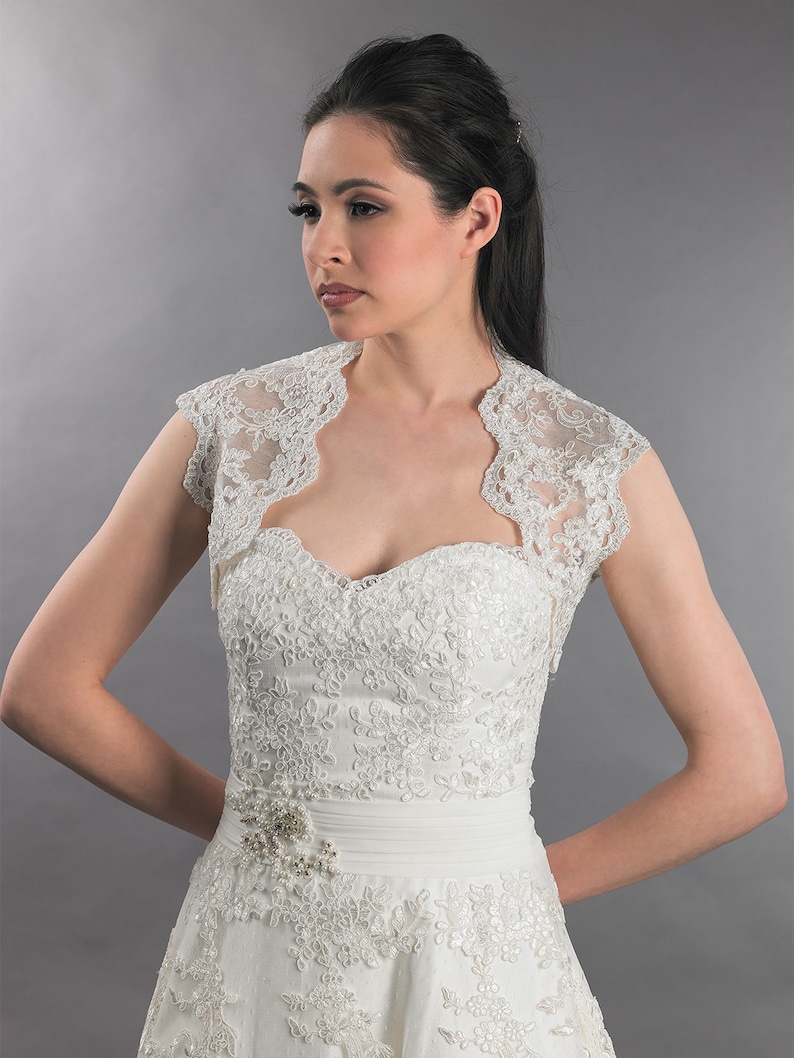May include: A white lace bridal bolero with short sleeves and a scoop neckline. The bolero is made of delicate lace and has a scalloped edge.