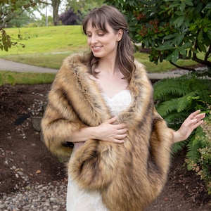 May include: A brown faux fur shawl with a long, flowing design. The shawl is worn by a person in a white dress, standing in a garden setting.