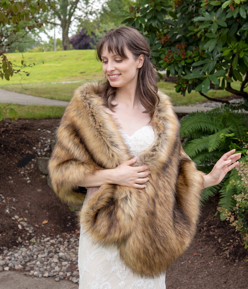 May include: A brown faux fur shawl with a long, flowing design. The shawl is worn by a person in a white dress, standing in a garden setting.