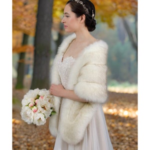May include: A white faux fur bridal wrap with a long, flowing design. The wrap is worn over a white wedding dress and is paired with a bouquet of white peonies.