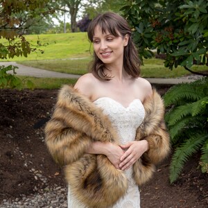 May include: A woman wearing a white strapless wedding dress with a brown faux fur shawl. She is standing in a garden setting.