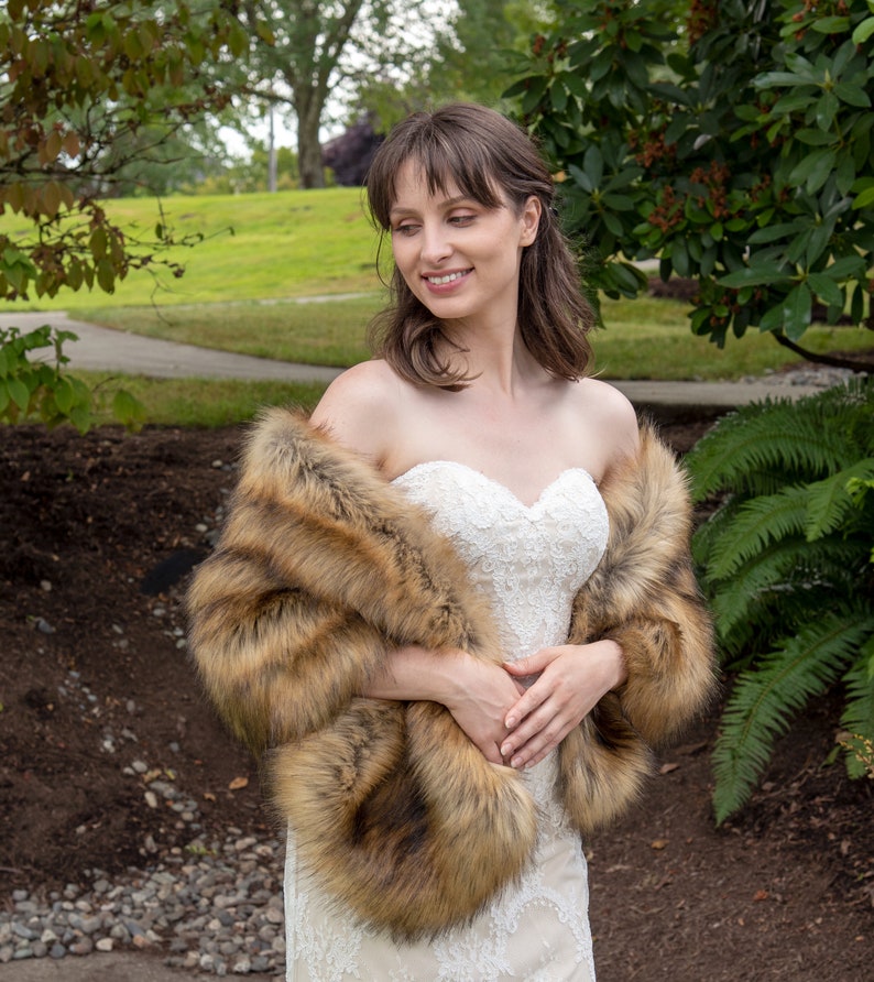 May include: A woman wearing a white strapless wedding dress with a brown faux fur shawl. She is standing in a garden setting.