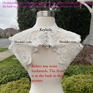 May include: A white lace bolero with a keyhole neckline. The bolero is worn backwards, with the front of the bolero at the back of the mannequin. The text "Bolero was worn backwards. The front is at the back in this picture." is written below the bolero.