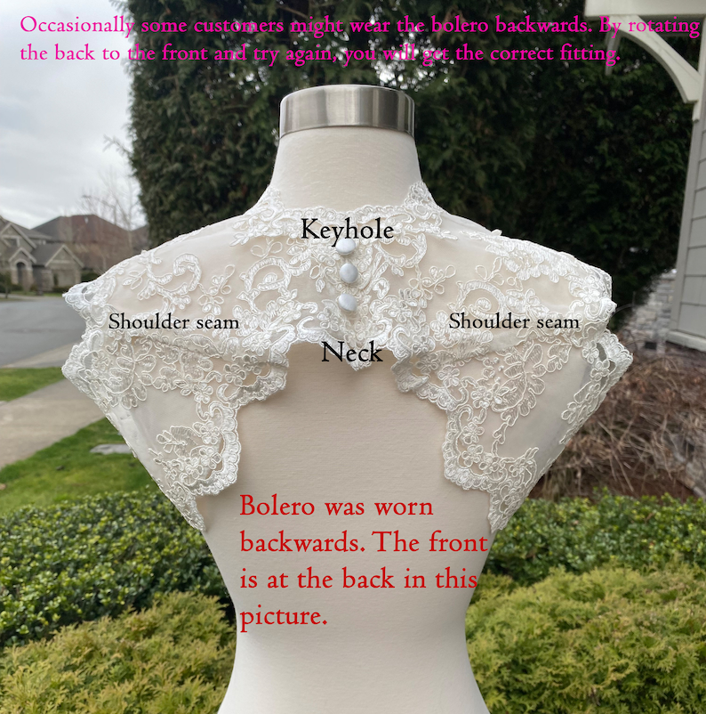 May include: A white lace bolero with a keyhole neckline. The bolero is worn backwards, with the front of the bolero at the back of the mannequin. The text "Bolero was worn backwards. The front is at the back in this picture." is written below the bolero.