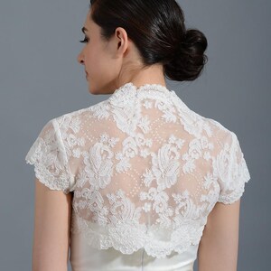 K&ouml;nnte beinhalten: Wei&szlig;er Brautbolero aus Spitze mit kurzen &Auml;rmeln und einem gewellten Saum. Der Bolero ist aus zarter Spitze mit Blumenmuster.