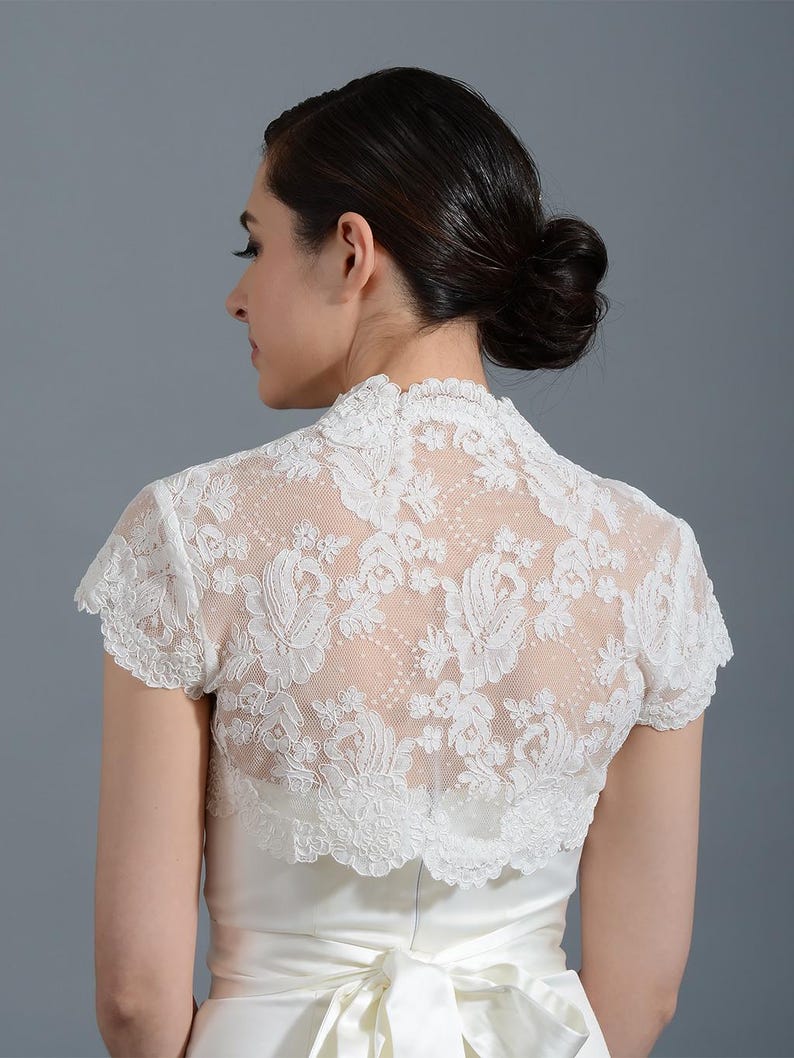 K&ouml;nnte beinhalten: Wei&szlig;er Brautbolero aus Spitze mit kurzen &Auml;rmeln und einem gewellten Saum. Der Bolero ist aus zarter Spitze mit Blumenmuster.