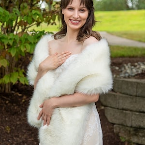 May include: A white faux fur shawl with dark brown tips. The shawl is draped around the shoulders of a person wearing a white dress.