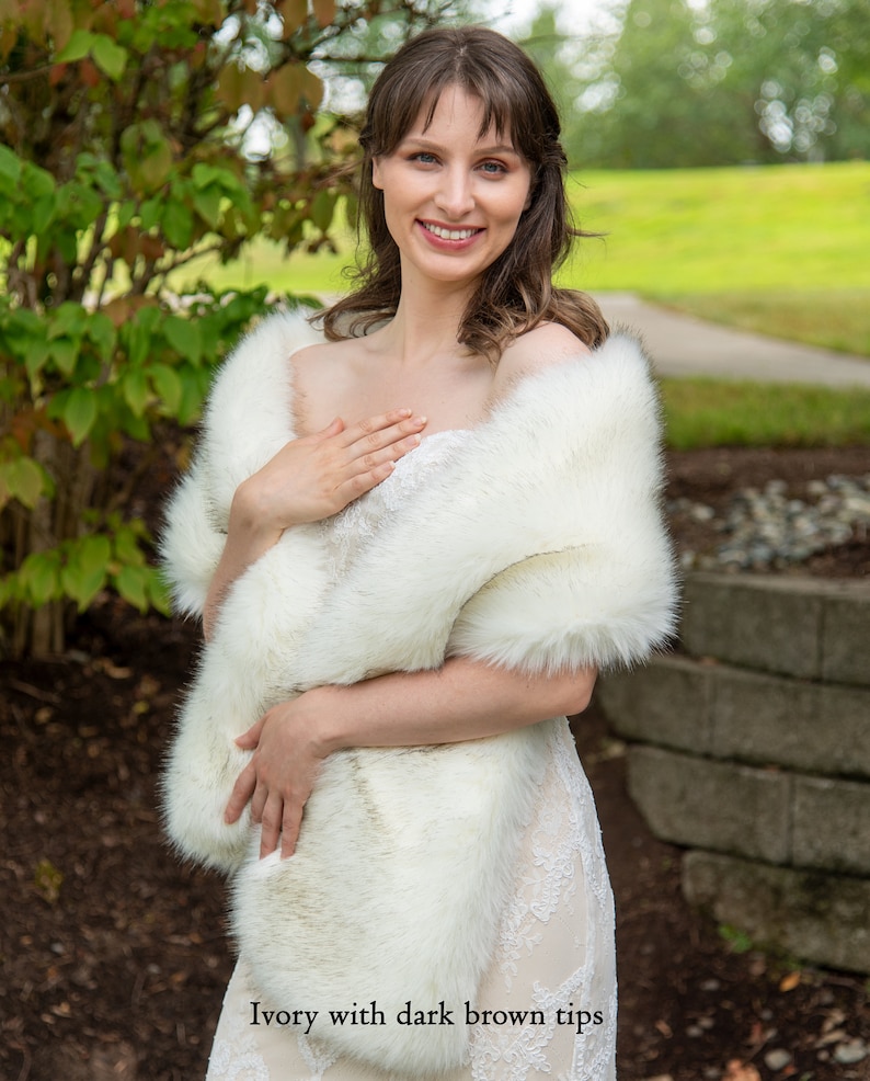 May include: A white faux fur shawl with dark brown tips. The shawl is draped around the shoulders of a person wearing a white dress.