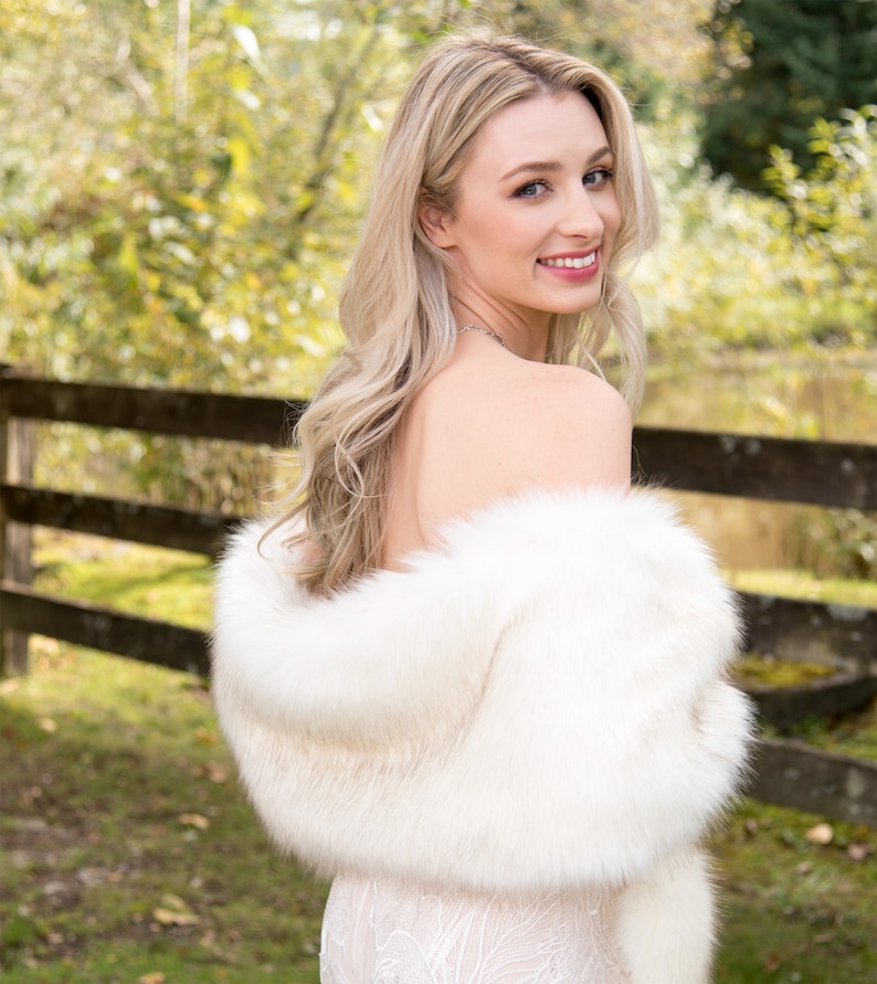 May include: A woman wearing a white faux fur shawl over a white lace dress. She is standing in front of a wooden fence with a green grassy background.