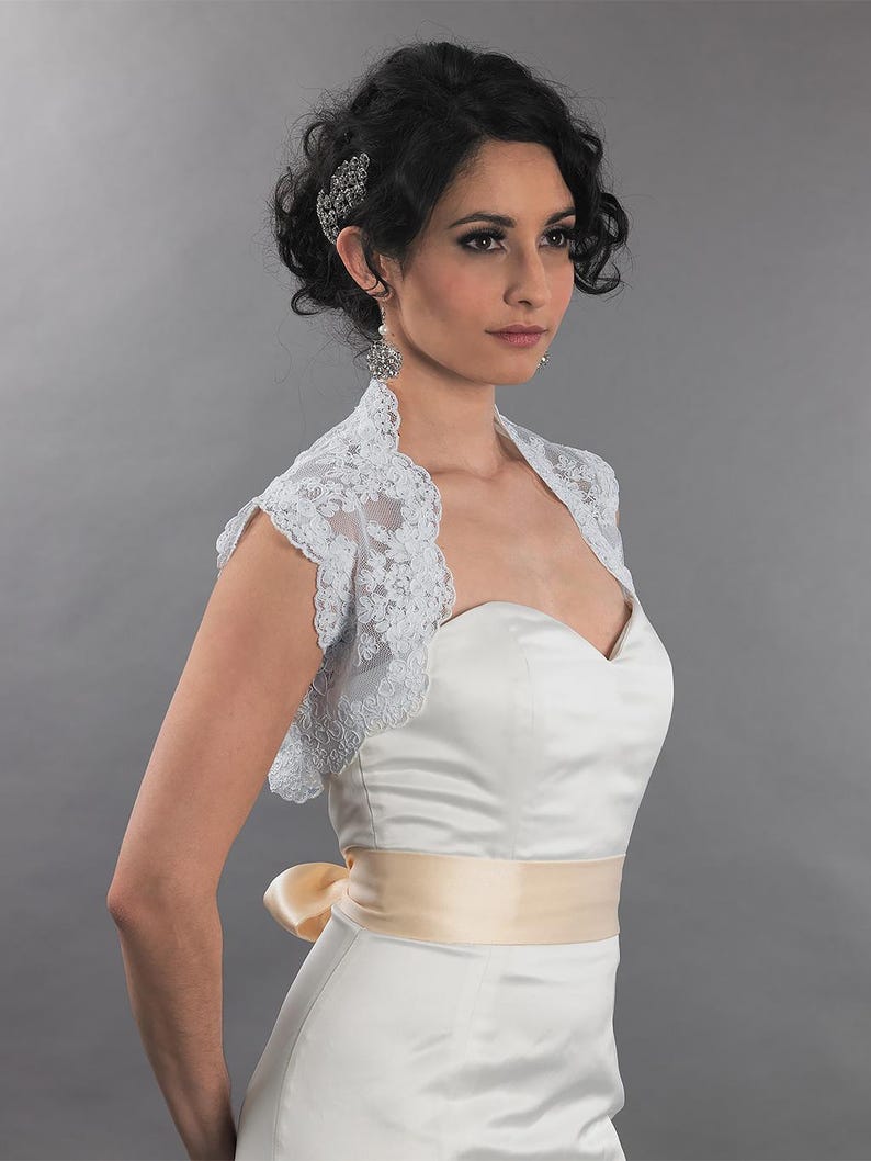 May include: A white satin wedding dress with a sweetheart neckline and a pale yellow sash. A delicate, light blue lace bolero covers the shoulders. The model is wearing a jeweled hair clip and earrings.