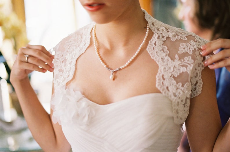 May include: A bride wearing a white lace wedding dress with a pearl necklace and a single pearl pendant.