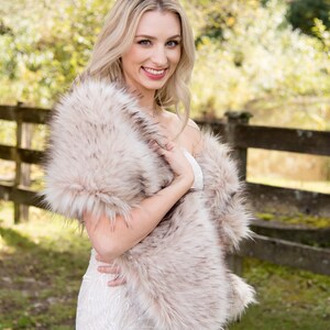 May include: A woman wearing a white dress with a faux fur shawl in a light brown color. The shawl is draped around her shoulders and she is holding it in front of her. She is standing in front of a wooden fence with green grass and trees in the background.