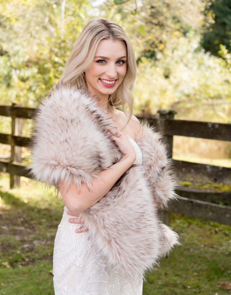 May include: A woman wearing a white dress with a faux fur shawl in a light brown color. The shawl is draped around her shoulders and she is holding it in front of her. She is standing in front of a wooden fence with green grass and trees in the background.