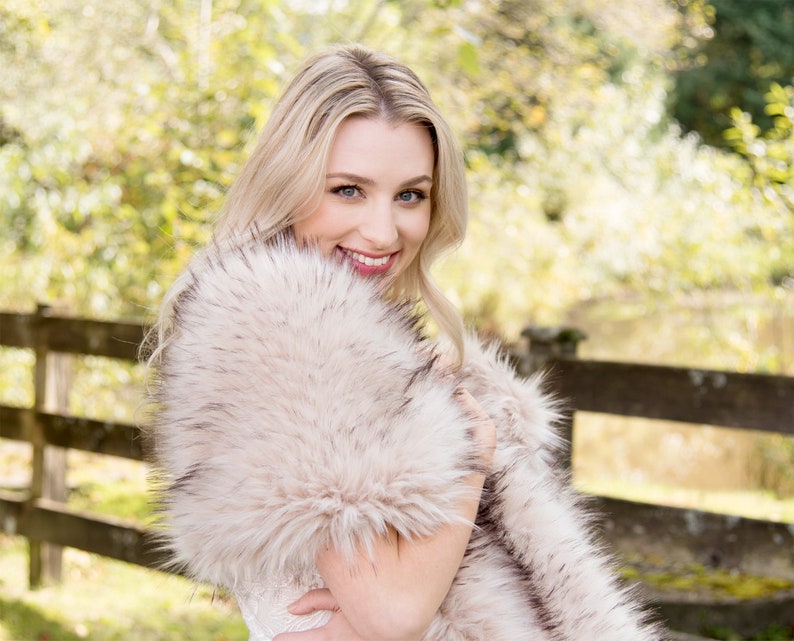 May include: A woman wearing a white dress with a faux fur shawl. The shawl is a light brown color with black tips. The woman is smiling and looking at the camera.
