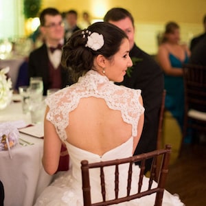 May include: A bride wearing a white lace wedding dress with a low back and a white flower in her hair. She is sitting in a wooden chair and smiling.