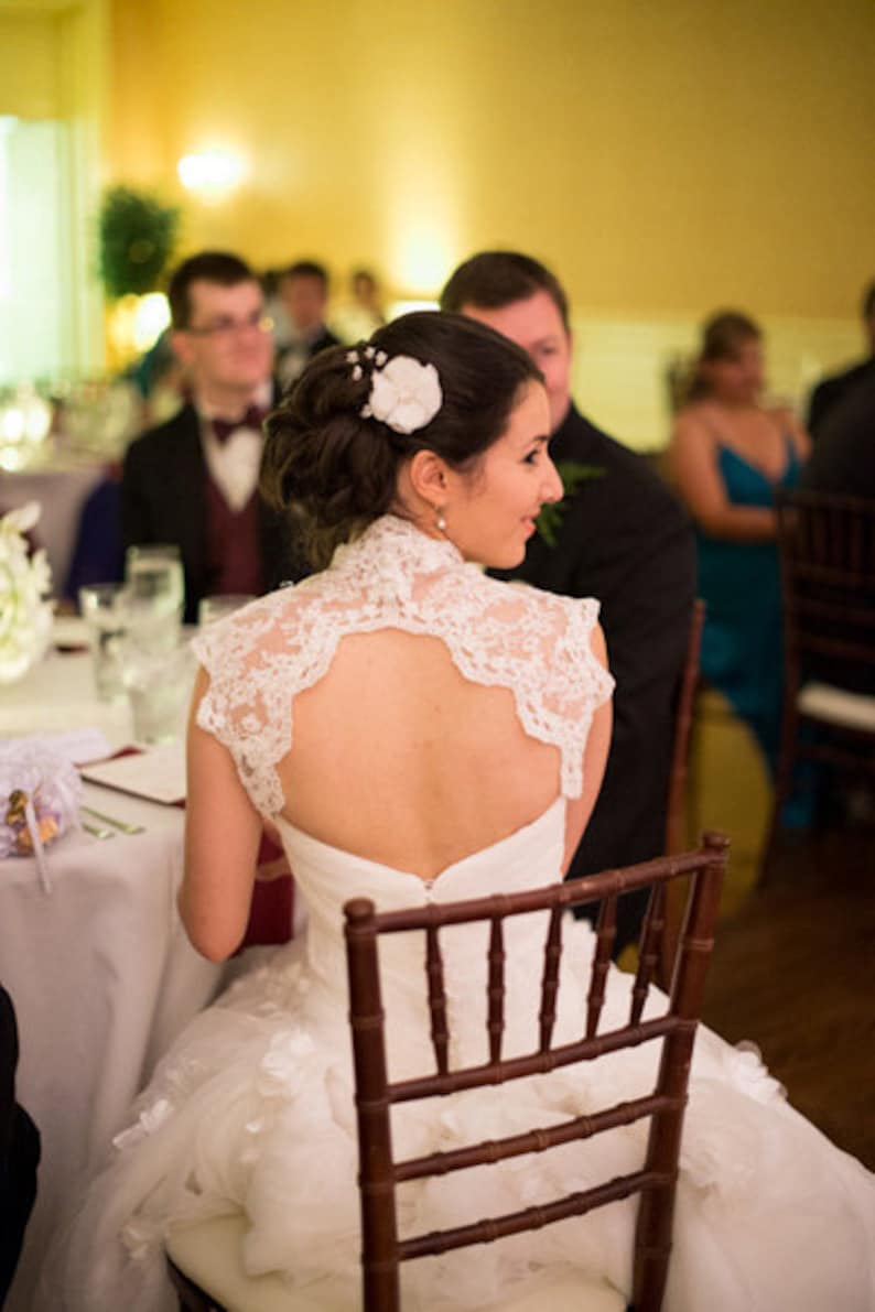May include: A bride wearing a white lace wedding dress with a low back and a white flower in her hair. She is sitting in a wooden chair and smiling.