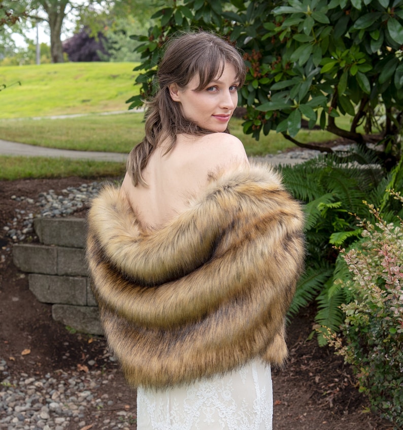 May include: A brown faux fur shawl with a long, flowing design. The shawl is draped over the shoulders of a person standing in a garden.