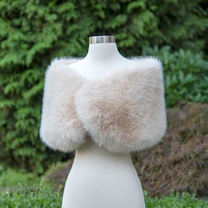 May include: A cream-coloured faux fur shawl with a soft, fluffy texture. The shawl is draped over a white mannequin.