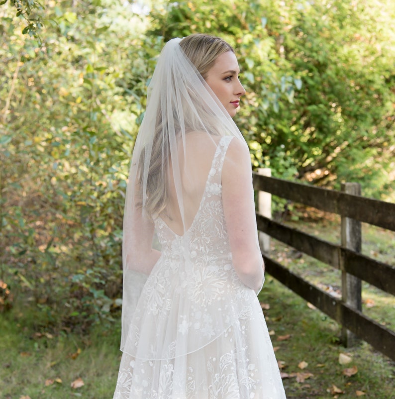 May include: A white bridal veil with a single layer of tulle, worn by a person with long blonde hair. The veil is simple and elegant, with a scalloped edge.