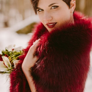 May include: A woman with short, dark red hair wears a burgundy faux fur shawl. She is holding a bouquet of white and red flowers.