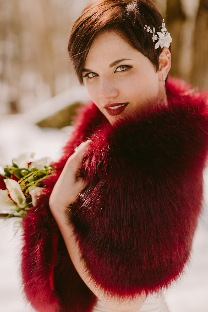 May include: A woman with short, dark red hair wears a burgundy faux fur shawl. She is holding a bouquet of white and red flowers.