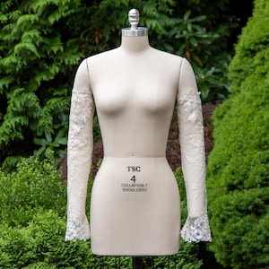 Könnte beinhalten: Eine cremefarbene Schneiderpuppe mit langen, transparenten Ärmeln, die mit weißer floraler Spitze verziert sind. Auf der Schneiderpuppe steht "TSC 4 COLLAPSIBLE SHOULDERS". Der Hintergrund ist ein grüner Garten. Die Ärmel enden in einer ausgestellten Spitzenmanschette.