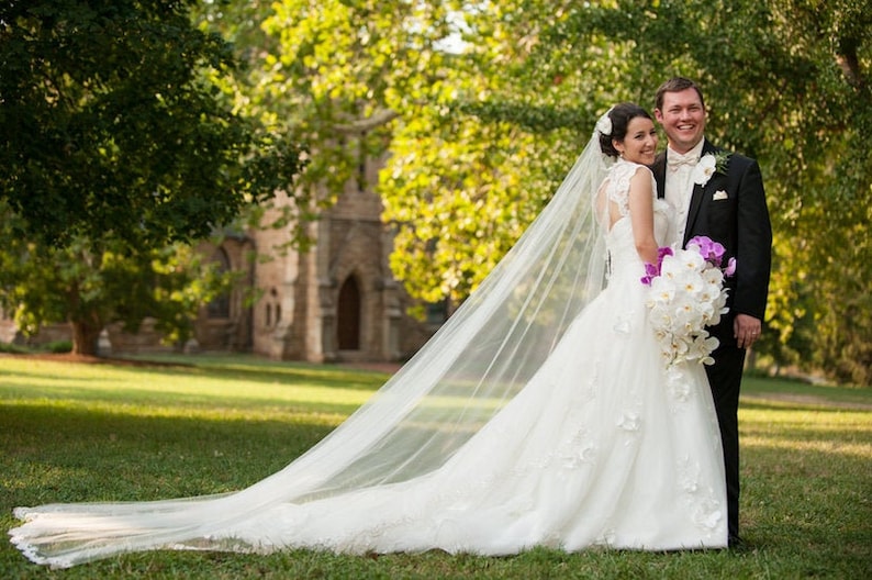 May include: A bride and groom pose for a wedding portrait in front of a stone building. The bride is wearing a white wedding dress with a long veil and a bouquet of white and purple orchids. The groom is wearing a black suit and a white shirt with a bow tie.