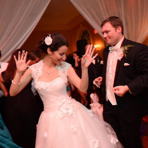 May include: A bride and groom dance at their wedding reception. The bride is wearing a white wedding dress with a lace bodice and a white flower in her hair. The groom is wearing a black tuxedo with a white shirt and a black bow tie. They are surrounded by guests who are dancing and celebrating.