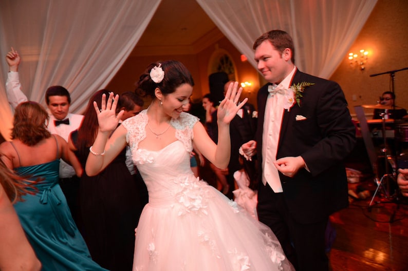 May include: A bride and groom dance at their wedding reception. The bride is wearing a white wedding dress with a lace bodice and a white flower in her hair. The groom is wearing a black tuxedo with a white shirt and a black bow tie. They are surrounded by guests who are dancing and celebrating.